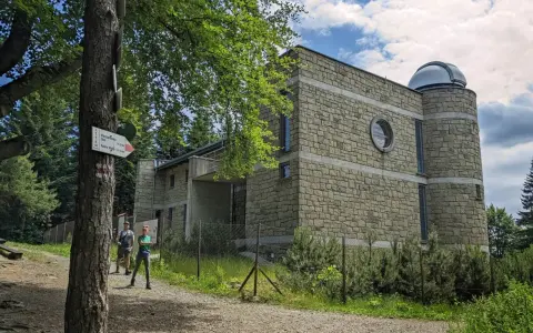 Kamienny budynek obserwatorium na szczycie Lubomir. Dwoje turystów podziwia widok, a obok drzewa i szlak PTTK.