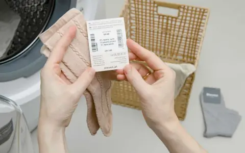 Ręce trzymają metkę z informacją, w czym prać wełnę merino, obok kosza i pralki.