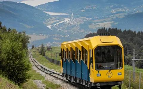 Kolejka linowo-terenowa na górę Żar z żółtym wagonikiem pnie się po torach, oferując widok na jezioro i dolinę.