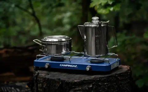Niebieska kuchenka turystyczna Campingaz z dwoma palnikami, na której stoją metalowy garnek i kawiarka, gotowa do przygotowania posiłku w lesie.